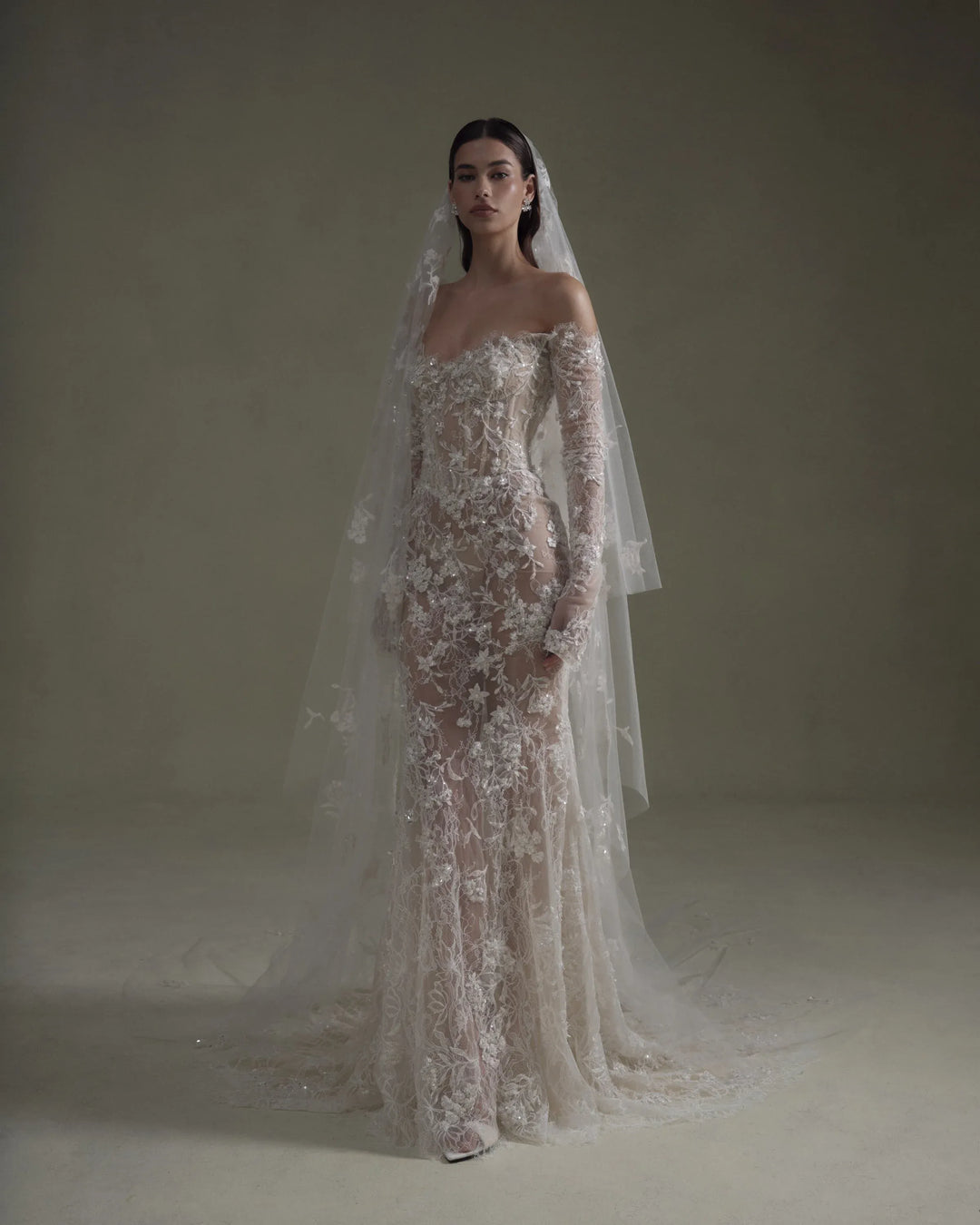 A model wearing an off-the-shoulder, floor-length mermaid-style wedding gown with intricate floral lace applique and a long sheer tulle veil, posed against a neutral, warm-toned studio backdrop.