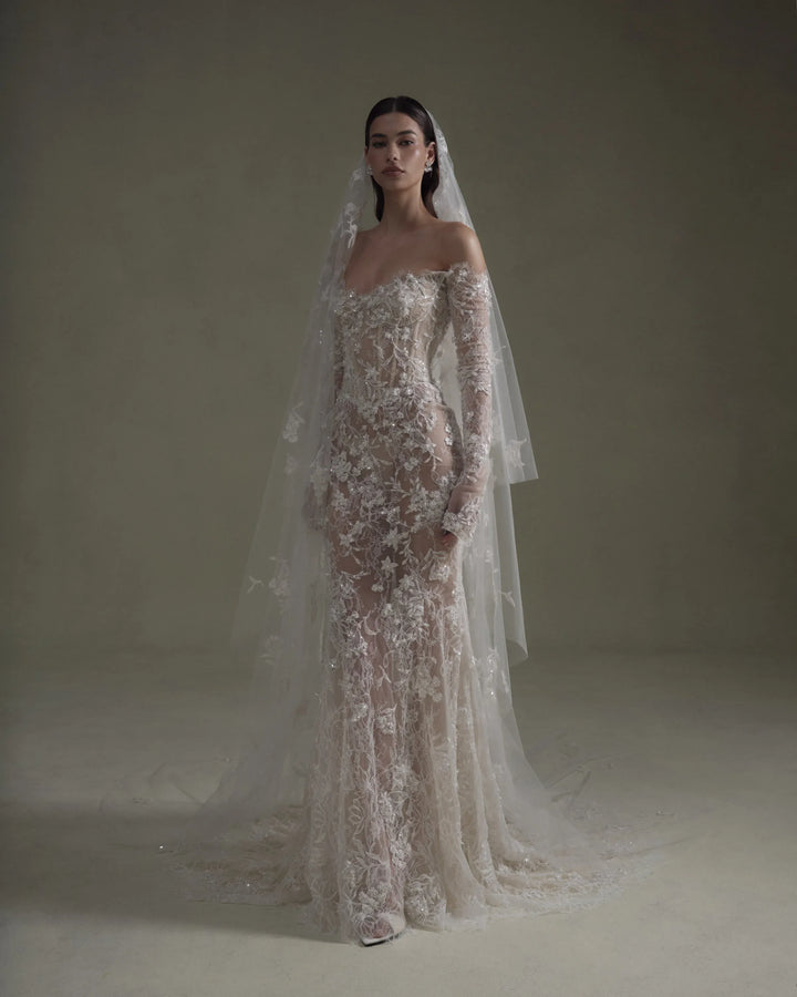 A model wearing an off-the-shoulder, floor-length mermaid-style wedding gown with intricate floral lace applique and a long sheer tulle veil, posed against a neutral, warm-toned studio backdrop.