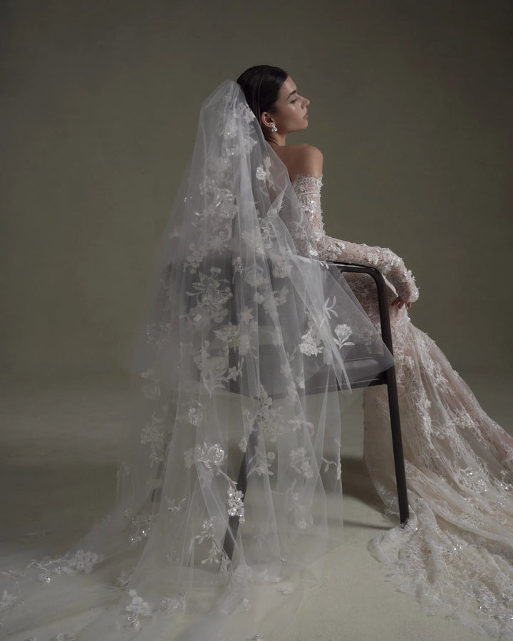 A woman in a bridal gown sits on a dark chair, shown from the back and side. She wears a long sheer tulle veil with floral lace applique and a voluminous embroidered wedding dress in a pale blush or ivory tone against a muted taupe studio background.