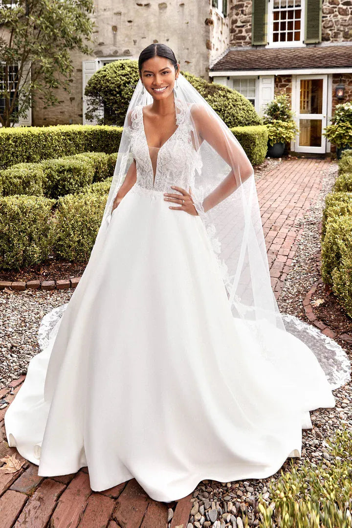 A woman wearing a white ballgown wedding dress with a long sheer veil stands outdoors on a brick path in front of a stone house and trimmed hedges.