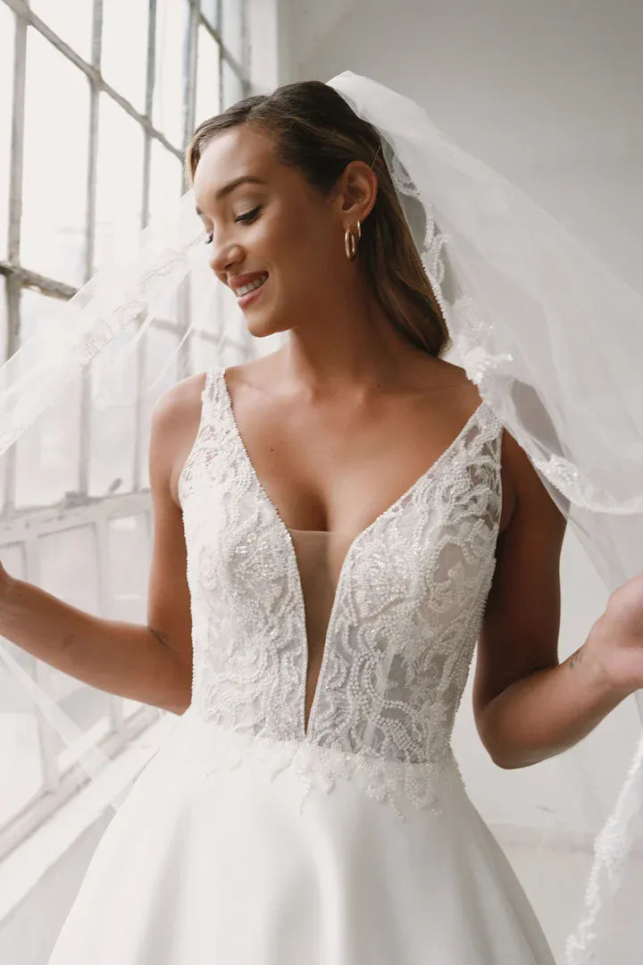 A woman wearing a white lace wedding dress with a deep V-neckline and a sheer veil, standing indoors near a bright window.