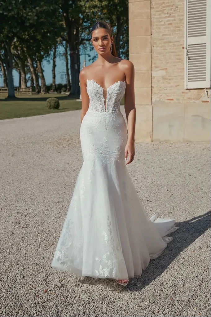 A woman stands outdoors in a white strapless mermaid-style wedding gown with a fitted lace bodice, sheer illusion neckline, and flared tulle skirt on a gravel courtyard beside a stone building.