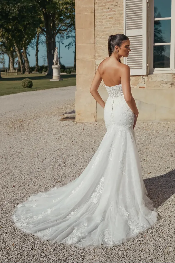 A woman wearing a strapless white mermaid-style wedding gown with floral lace applique and a long train, standing outdoors beside a stone building and gravel path.