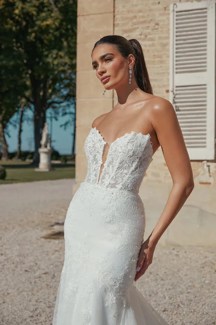A woman wearing a strapless white lace wedding gown with a sweetheart neckline and a fitted mermaid silhouette, standing outdoors in front of a beige stone building with shutters and trees in the background.