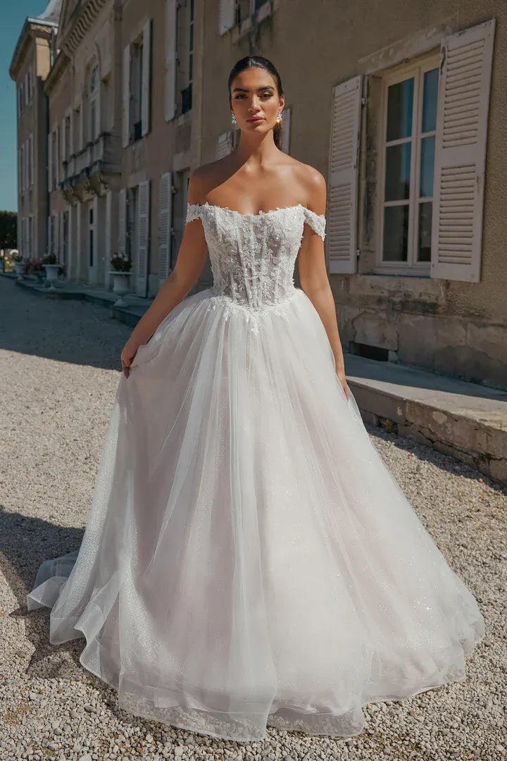 A woman modeling an off-the-shoulder A-line wedding gown with a lace-embroidered bodice and a voluminous tulle skirt, standing outdoors on a gravel path in front of a European-style building.