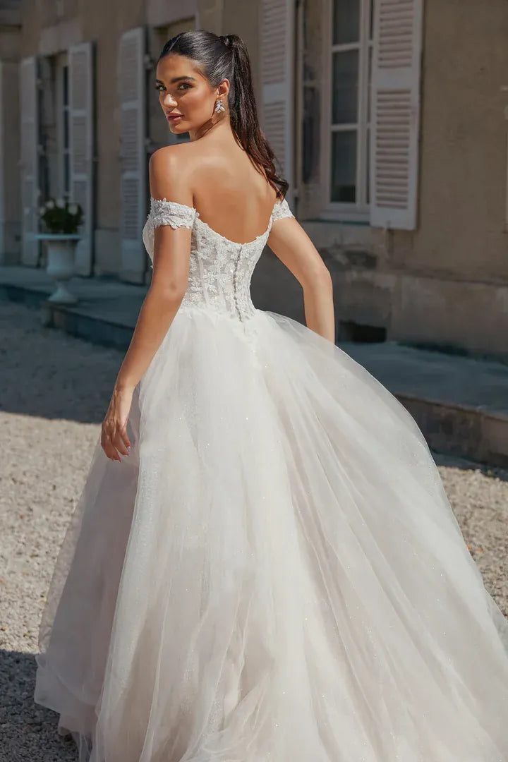 A woman wearing an off-the-shoulder ball gown wedding dress with a lace corset back and a full tulle skirt, standing outdoors in front of a building with shutters.