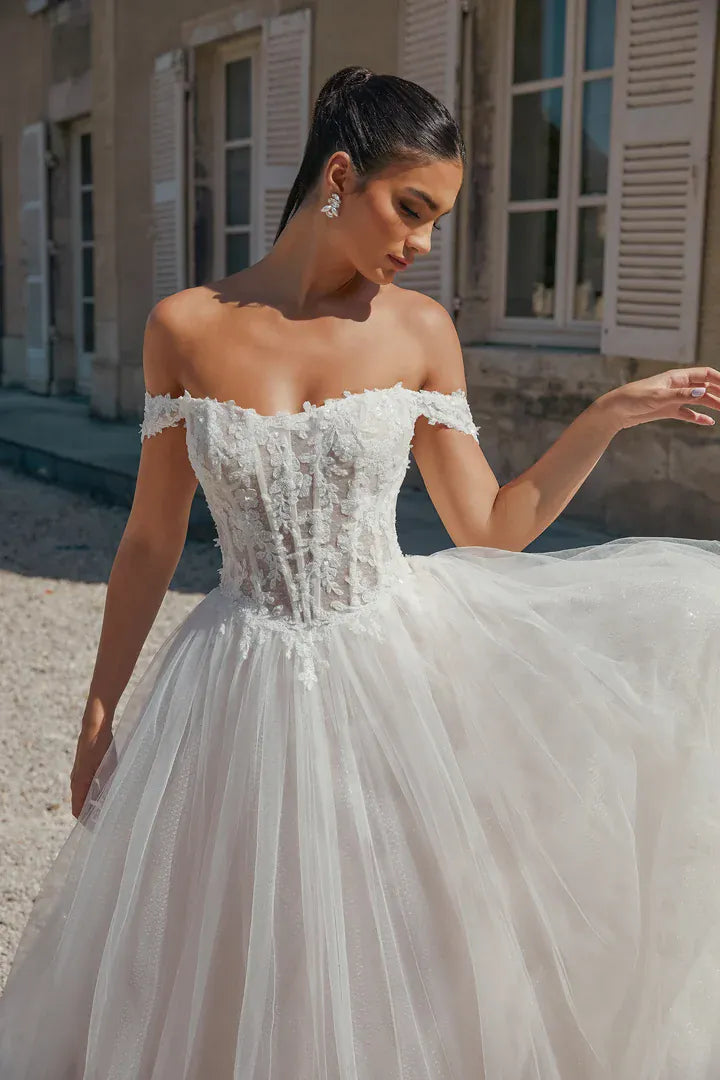 An off-the-shoulder white wedding gown with a lace-embellished corset bodice and a voluminous tulle skirt, worn by a model in front of a building with beige shutters.