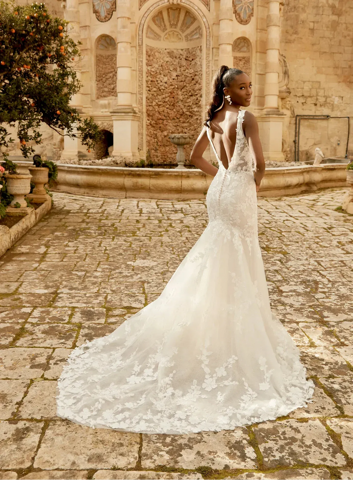 A woman wearing a white lace mermaid-style wedding gown with a deep V back and long train, standing in a stone courtyard in front of an ancient building.