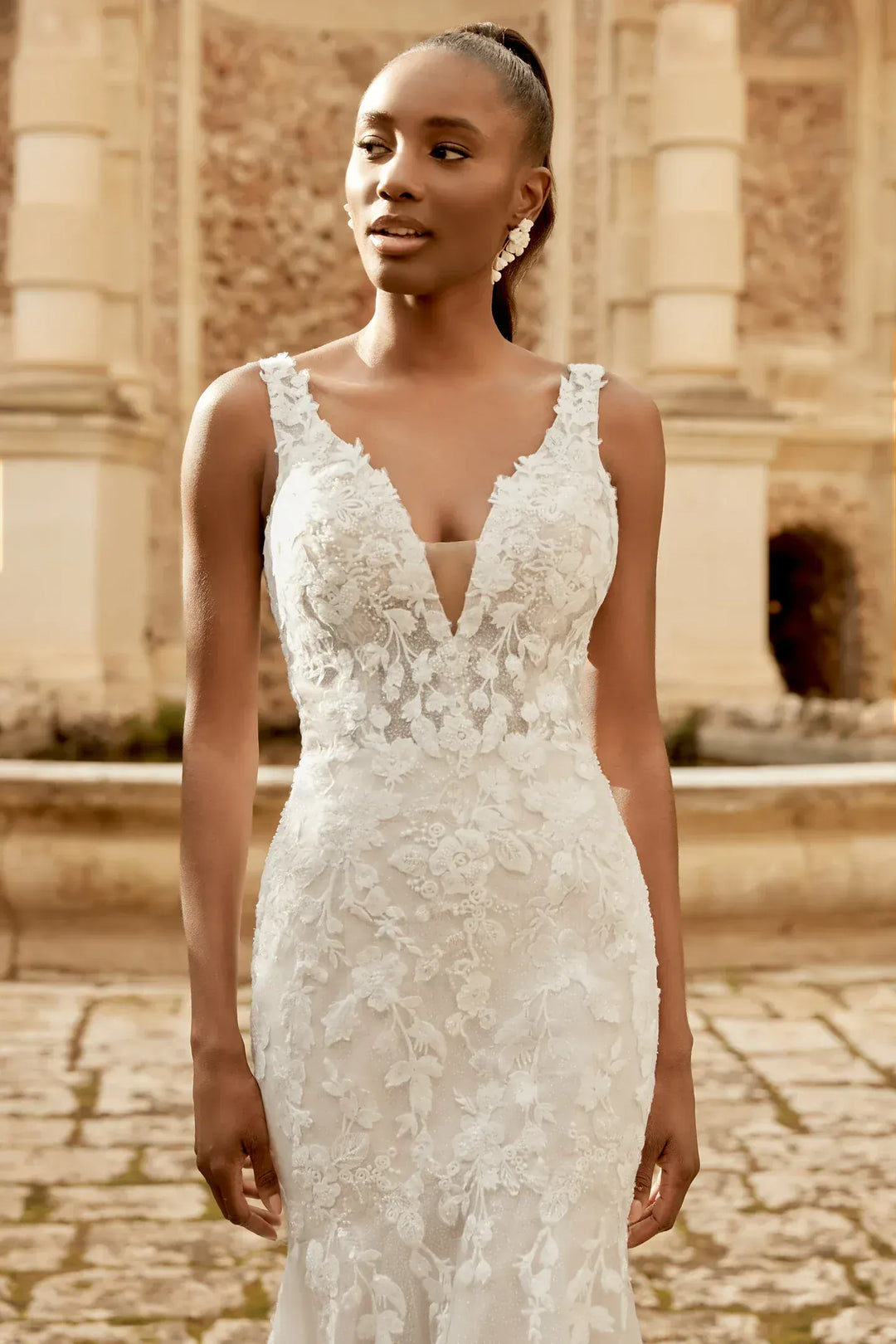 A woman wearing a fitted white lace wedding dress with a deep V-neckline and sleeveless straps, standing outdoors in front of a warm-toned stone building.