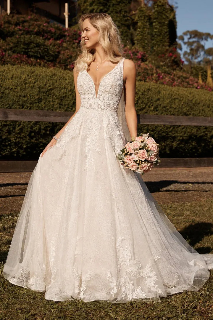 A woman wearing a white lace A-line wedding gown with a deep V-neckline and holding a pink bouquet, standing outdoors in front of a wooden fence and greenery.