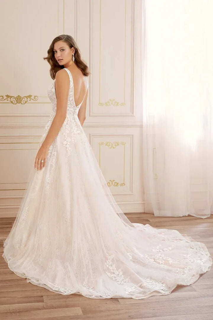 A woman modeling a white lace wedding gown with a deep V back and long train in an elegant cream-colored room with gold paneling and sheer curtains.