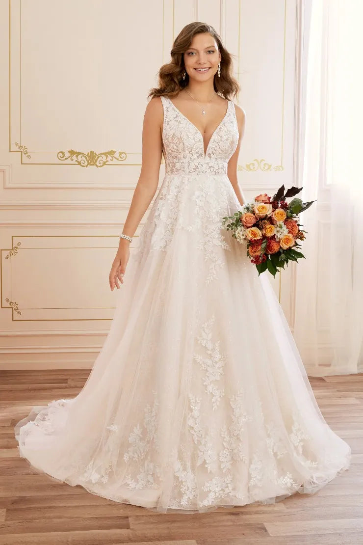 A woman modeling a sleeveless V-neck wedding gown with a fitted lace bodice and a full tulle skirt, standing indoors and holding a bouquet of orange, peach, and green flowers.