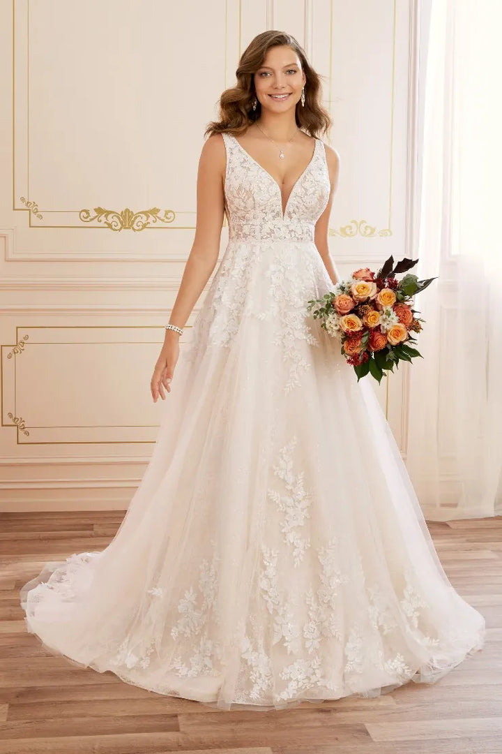 A woman modeling a sleeveless V-neck wedding gown with a fitted lace bodice and a full tulle skirt, standing indoors and holding a bouquet of orange, peach, and green flowers.