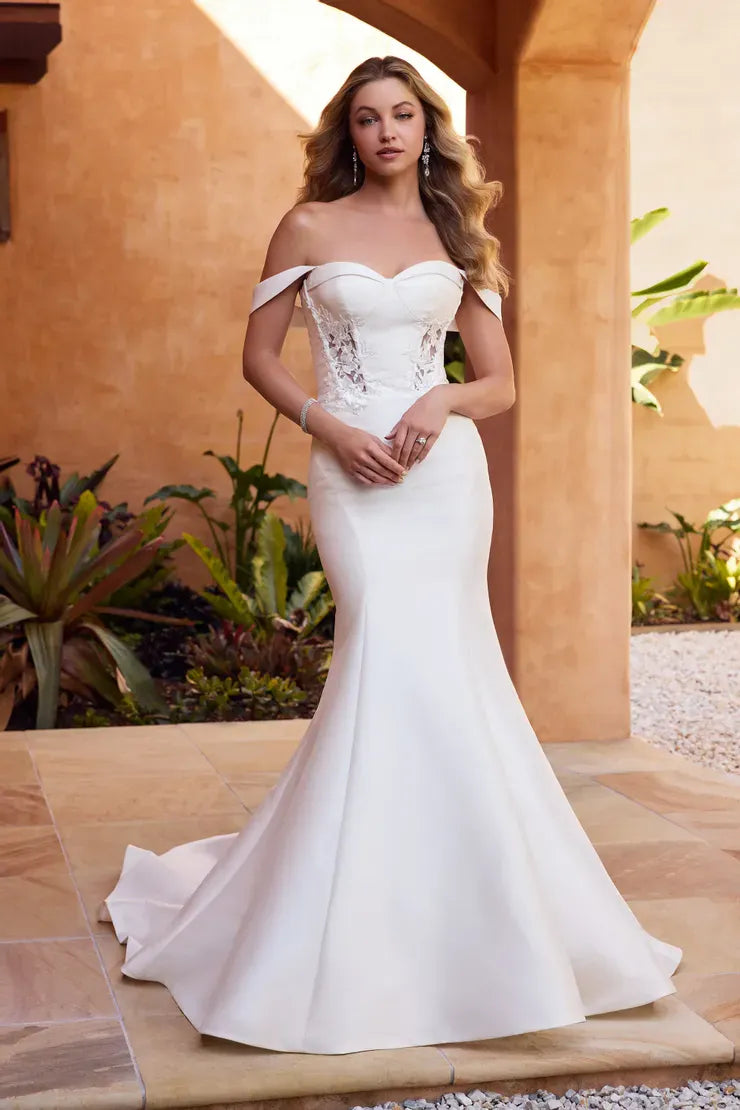 A woman modeling an off-the-shoulder white wedding gown with a fitted bodice, lace-up front, and flared mermaid skirt, standing in a warm-toned outdoor courtyard with stone columns and plants.