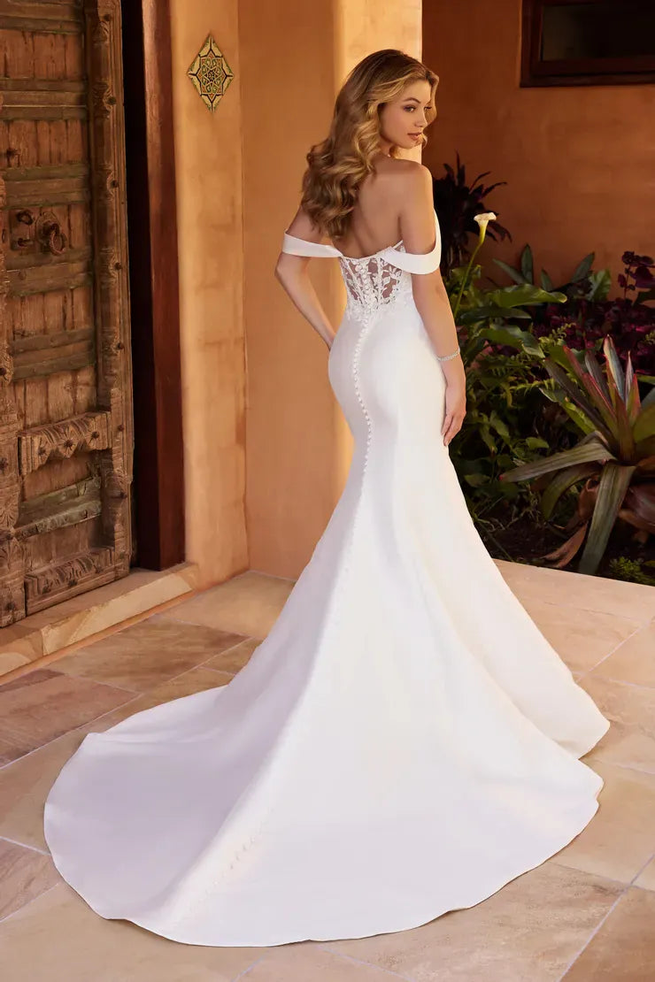 A woman wearing a white off-the-shoulder mermaid-style wedding gown with lace up the back, standing in a warm-toned indoor setting with a tiled floor, wooden door, and potted plants.