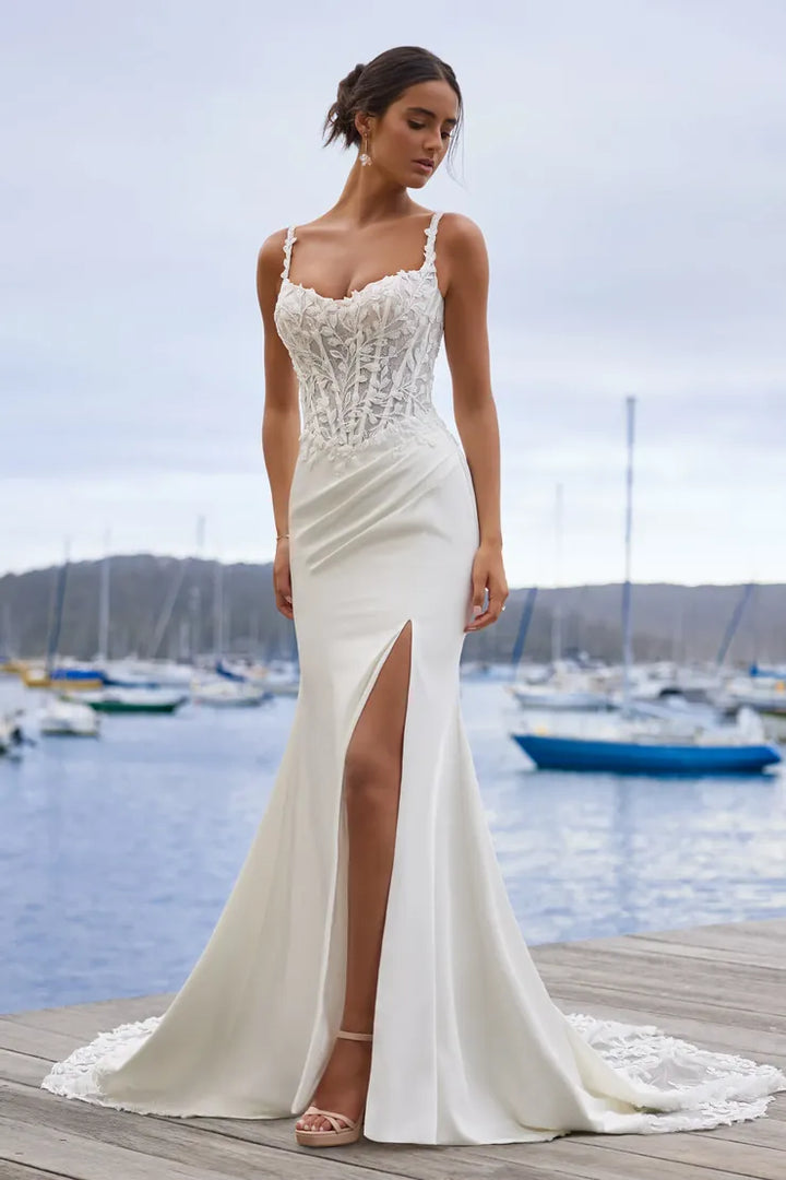 A woman wearing a sleeveless ivory wedding gown with a lace corset bodice, thin straps, and a high thigh slit, standing on a wooden dock with a harbor and boats in the background.