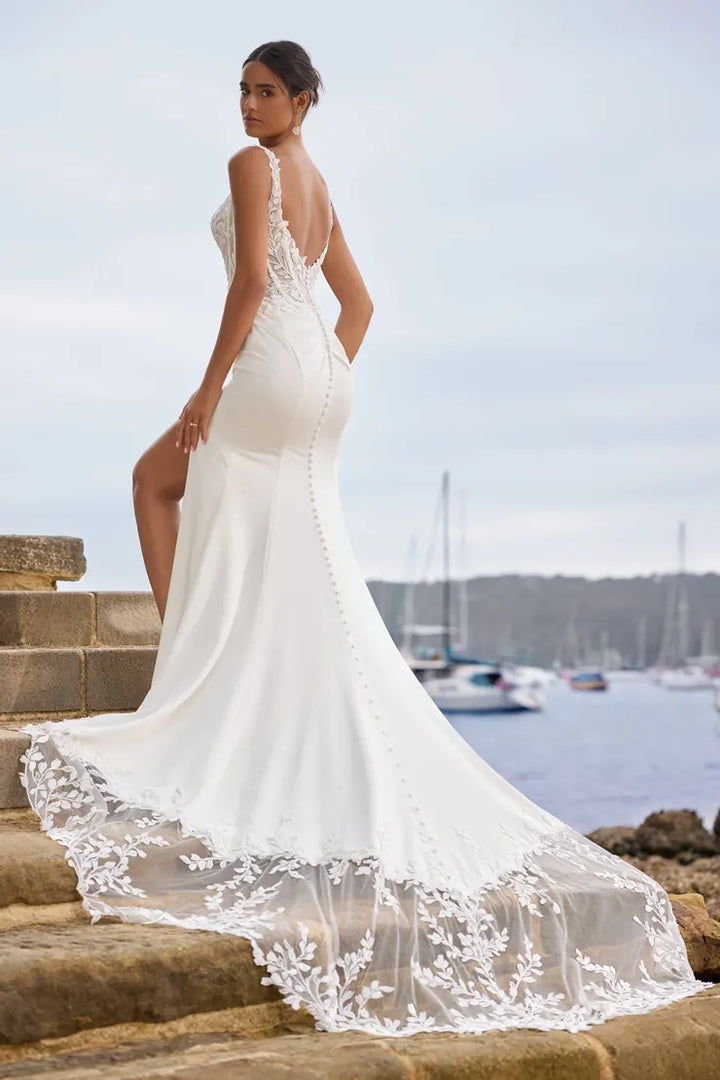 A woman modeling a white wedding gown from the back while standing on stone steps outdoors near the water, with boats and a cloudy sky in the background.
