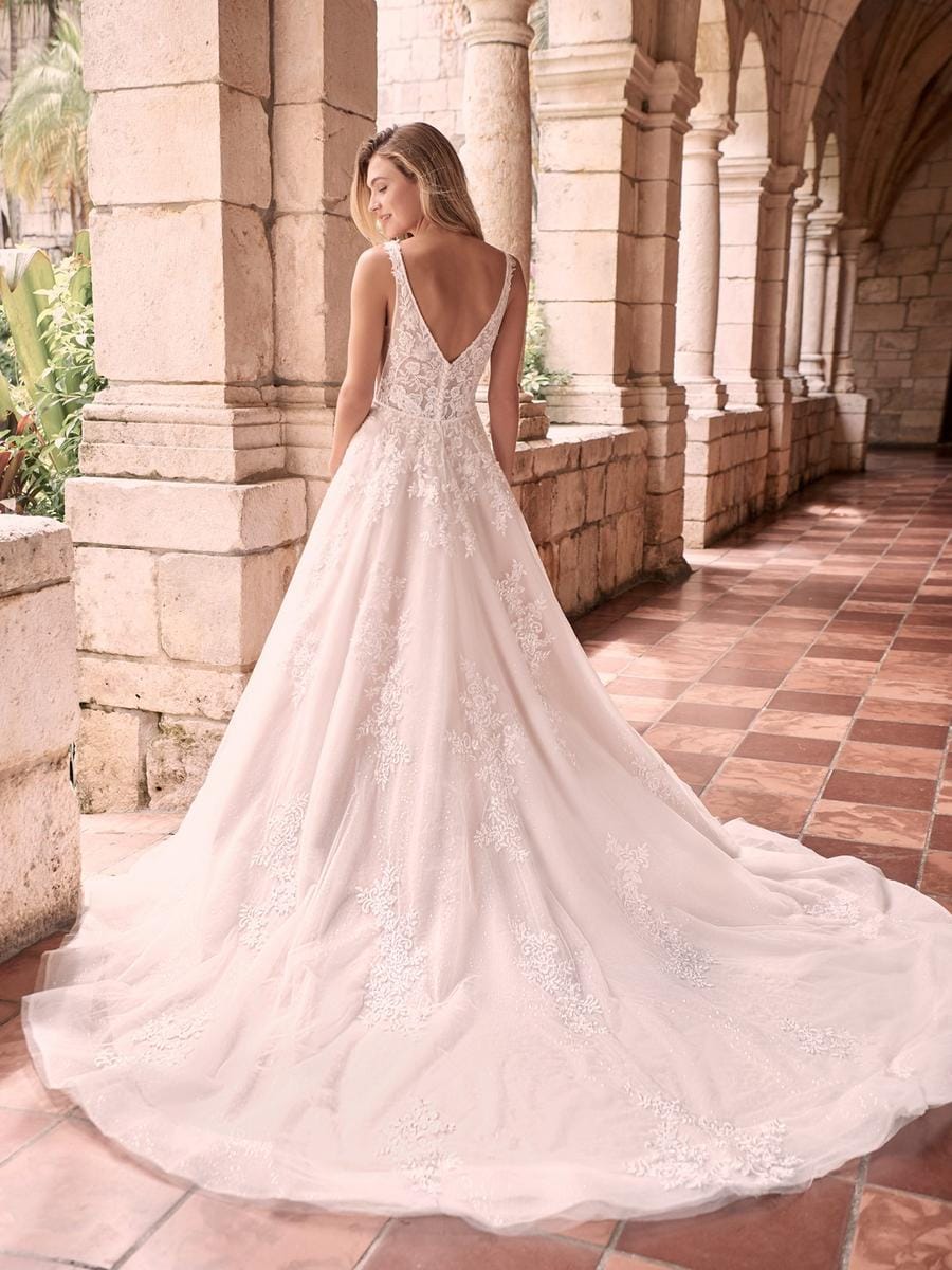 A woman wearing a bridal gown stands in a sunlit stone colonnade, photographed from the back. The dress has a deep V back, lace applique, and a full satin train spread across the tiled floor.