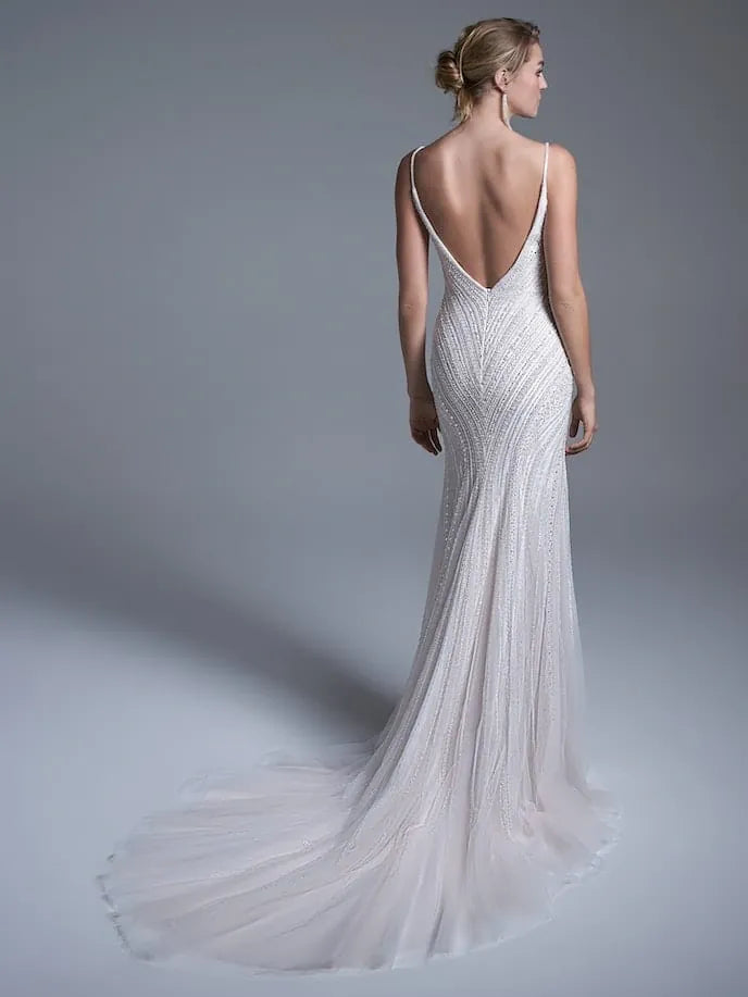 A woman modeling a sleeveless, backless white wedding gown from the back, showing a deep V-back and a long flowing tulle train against a smooth gray studio background.