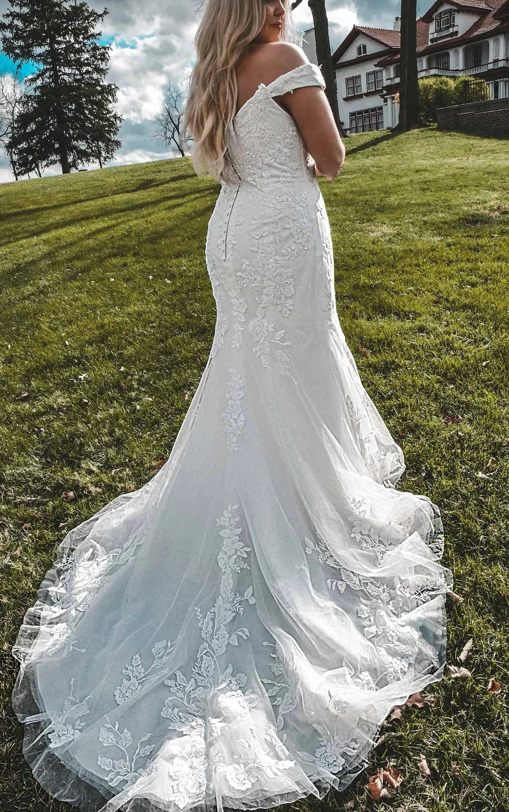 Woman in a white lace wedding dress standing outdoors with green grass and houses in the background