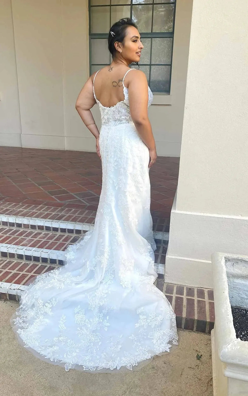 A woman wearing a white lace mermaid-style wedding gown with thin straps and a fitted silhouette, photographed outdoors on brick steps beside a beige building.