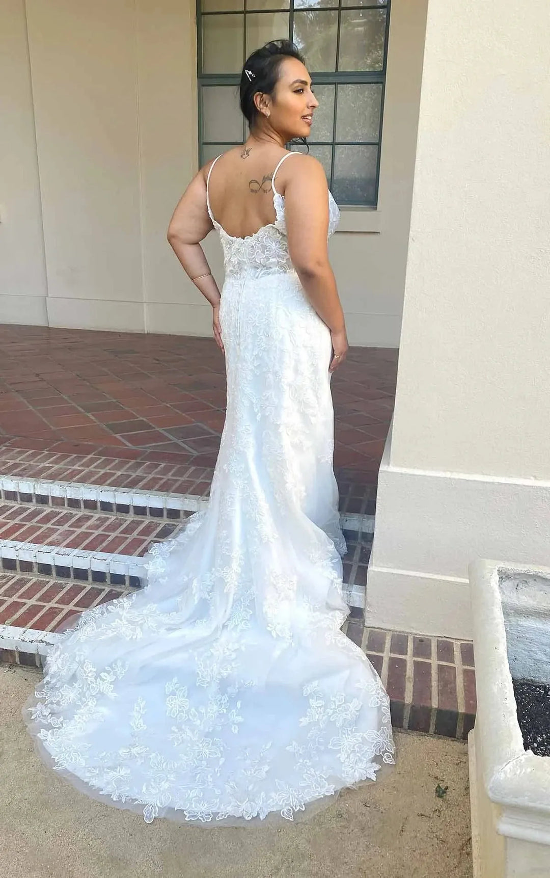 A woman wearing a white lace mermaid-style wedding gown with thin straps and a fitted silhouette, photographed outdoors on brick steps beside a beige building.