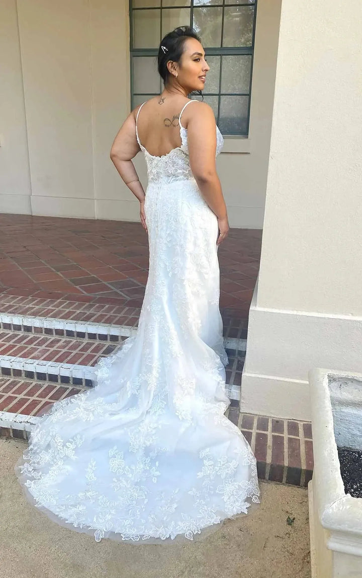 A woman wearing a white lace mermaid-style wedding gown with thin straps and a fitted silhouette, photographed outdoors on brick steps beside a beige building.