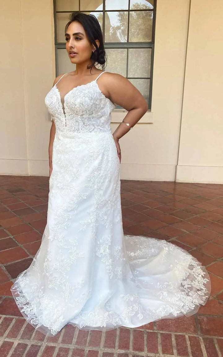 A woman modeling a white lace wedding gown with thin straps, a deep V-neckline, and a fitted mermaid silhouette, standing on a brick patio in front of a beige building wall.