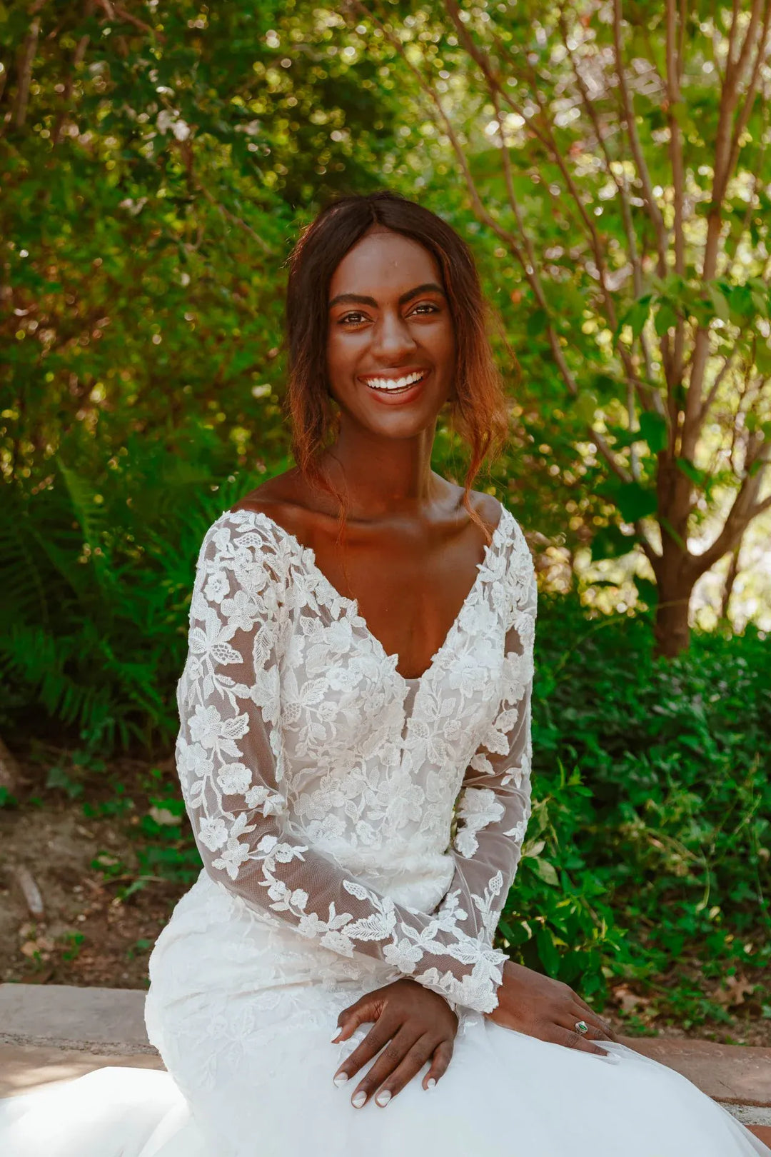 Woman in a white lace wedding dress sitting outdoors with greenery in the background