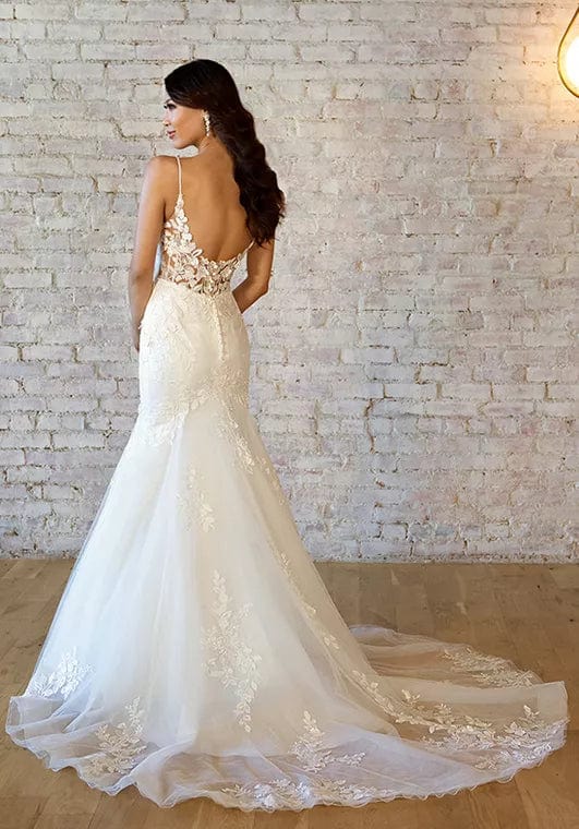 Woman wearing a white wedding dress with intricate detailing in front of a white brick wall.