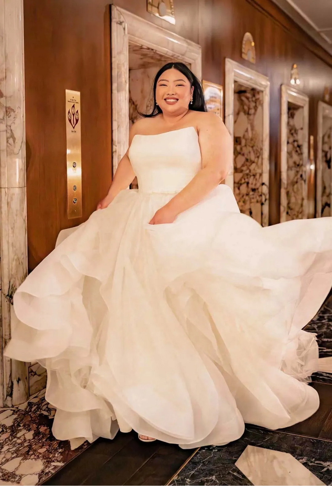Woman in a white wedding dress standing in a hallway with marble walls.