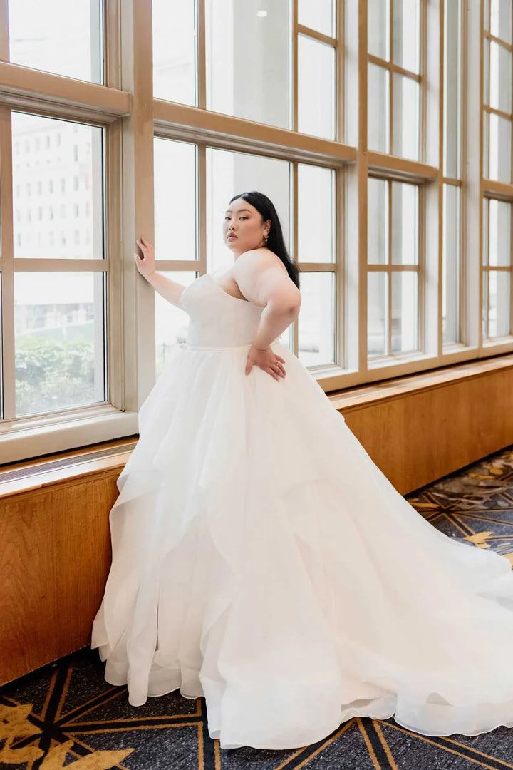 A woman wearing a strapless white tulle wedding gown stands by large wood-framed windows in a bright indoor setting with a patterned carpet visible.