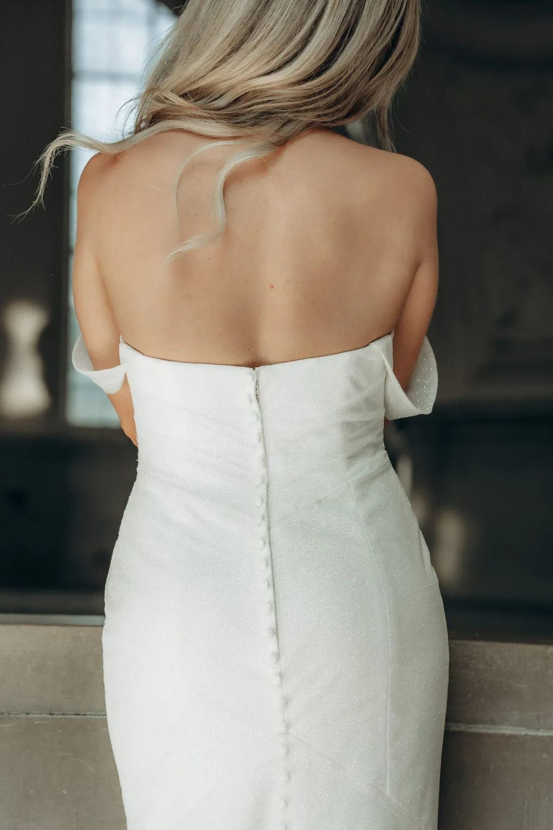 A woman shown from behind wearing a strapless white wedding dress with a fitted bodice and a central button closure, photographed indoors against a softly blurred dark background.