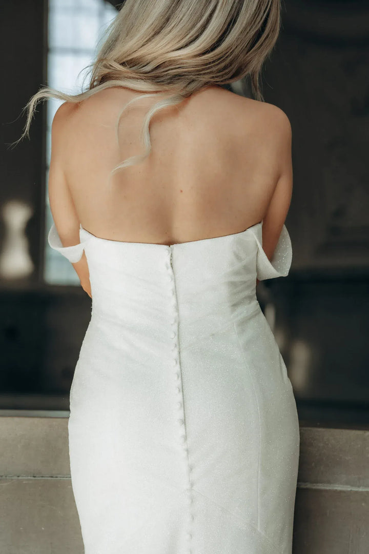 A woman shown from behind wearing a strapless white wedding dress with a fitted bodice and a central button closure, photographed indoors against a softly blurred dark background.