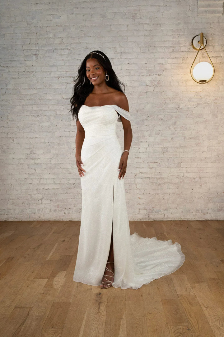 A woman modeling an off-the-shoulder white wedding gown with a thigh-high slit and a long flowing train, standing in a room with a light brick wall and wood floor.