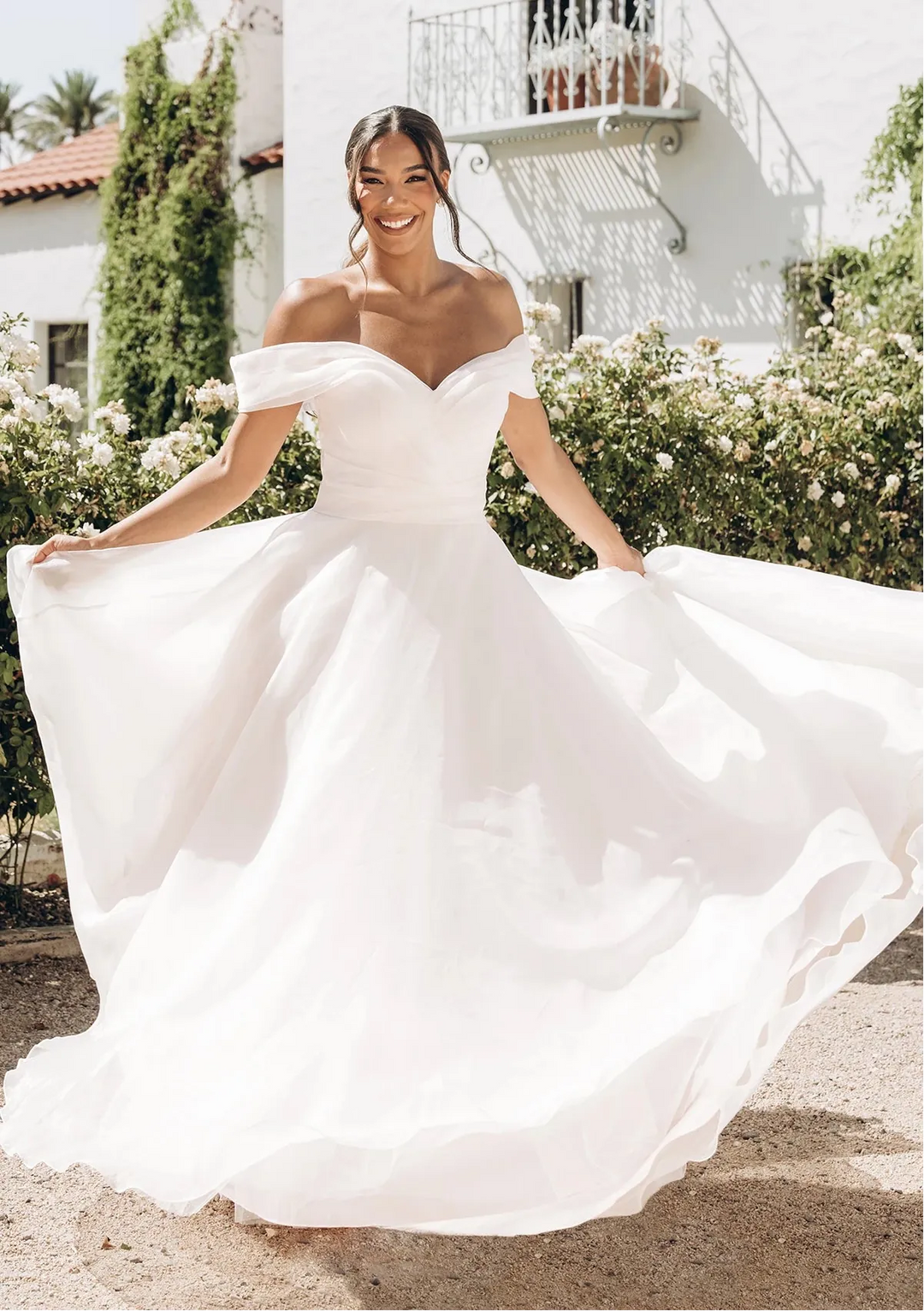 Woman in a white wedding dress standing outdoors with a building and greenery in the background