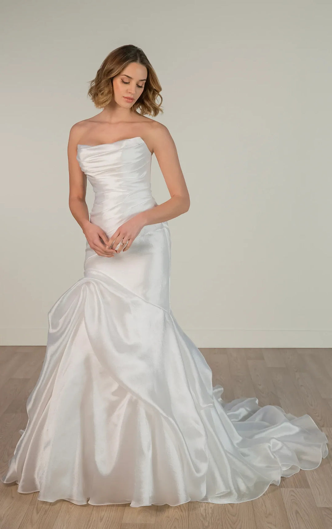 A woman modeling a strapless white satin mermaid-style wedding gown with a fitted bodice, flared skirt, and long train against a plain light beige wall and wood floor.