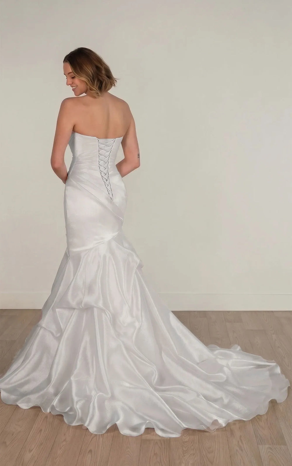 A woman wearing a strapless white satin mermaid-style wedding gown with a corseted back and full train, photographed in a minimal indoor room with light wood flooring and a plain off-white wall.