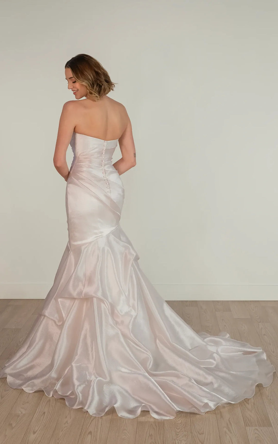 A woman wearing a strapless satin mermaid-style wedding gown, photographed from the back in a bright indoor room with a light wall and wood floor.