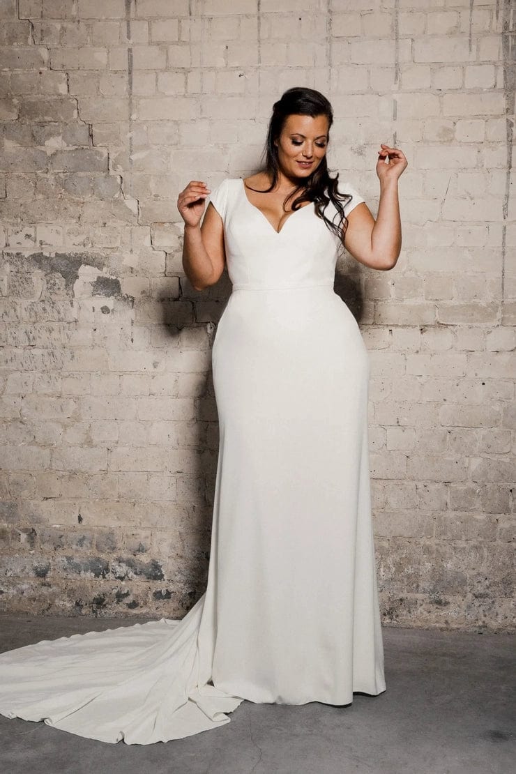 A woman modeling a floor-length white wedding gown with a deep V-neckline and a short train, standing against a textured brick wall.