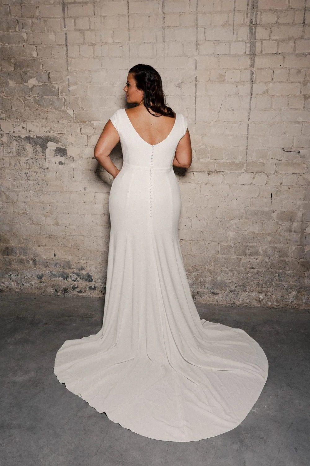 A woman stands with her back to the camera wearing a fitted white wedding gown with a deep V back and a long train, posed against a textured brick wall.