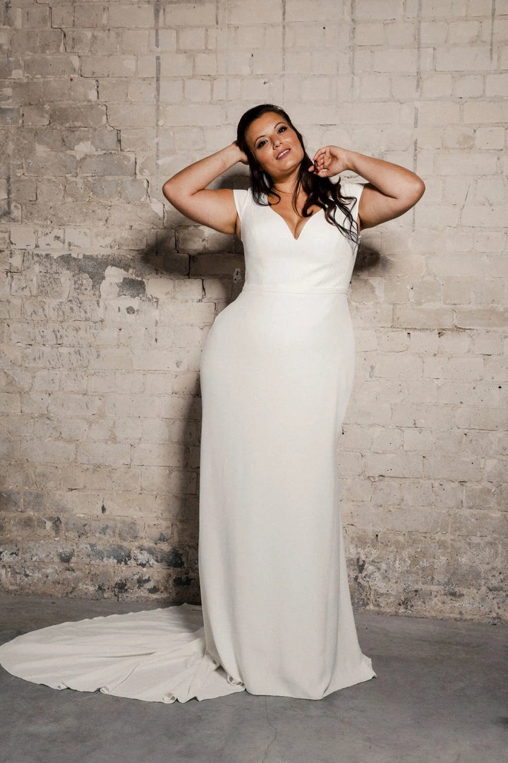 A woman modeling a floor-length white wedding dress with a deep V-neckline and sleeveless cap sleeves, standing against a textured light gray brick wall with a concrete floor.