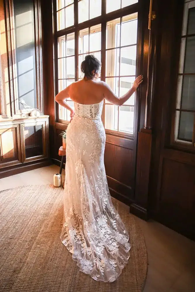 A woman viewed from behind wearing a strapless lace wedding gown with a fitted bodice and full train, standing indoors near large dark wood windows.