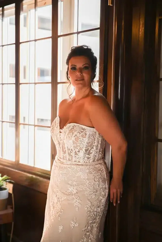 A woman wearing a strapless ivory lace wedding gown stands indoors beside a large window and dark wooden door frame.