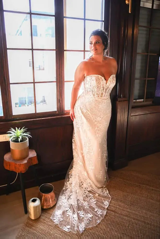 A woman wearing a strapless lace wedding gown with a sweetheart neckline and fitted corset-style bodice, standing indoors near a large window with dark wood paneling. A small side table with a plant and copper pots are visible on the floor.