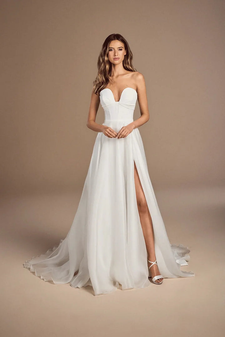 A woman modeling a strapless white wedding gown with a sweetheart neckline, fitted bodice, high front slit, and a flowing train against a warm beige studio backdrop.