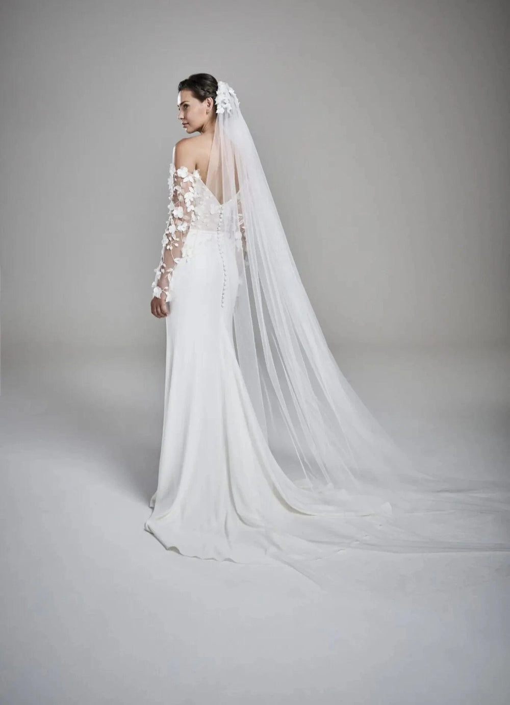 A woman modeling a white off-the-shoulder wedding gown with floral lace sleeves, a long sheer veil, and a fitted silhouette, photographed against a soft light gray studio background.