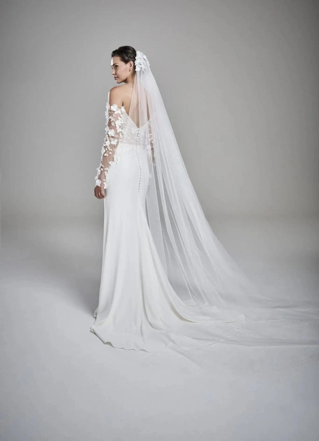 A woman modeling a white off-the-shoulder wedding gown with floral lace sleeves, a long sheer veil, and a fitted silhouette, photographed against a soft light gray studio background.