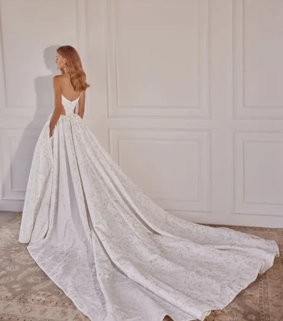 A woman wearing a white lace bridal gown with a long train, photographed indoors against a cream paneled wall.