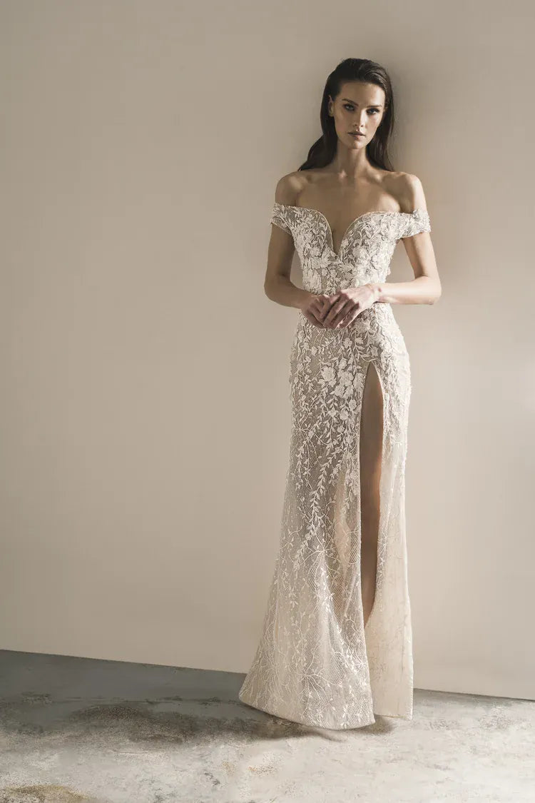 A woman modeling an off-the-shoulder, floor-length lace gown with a thigh-high slit, standing against a plain beige wall.